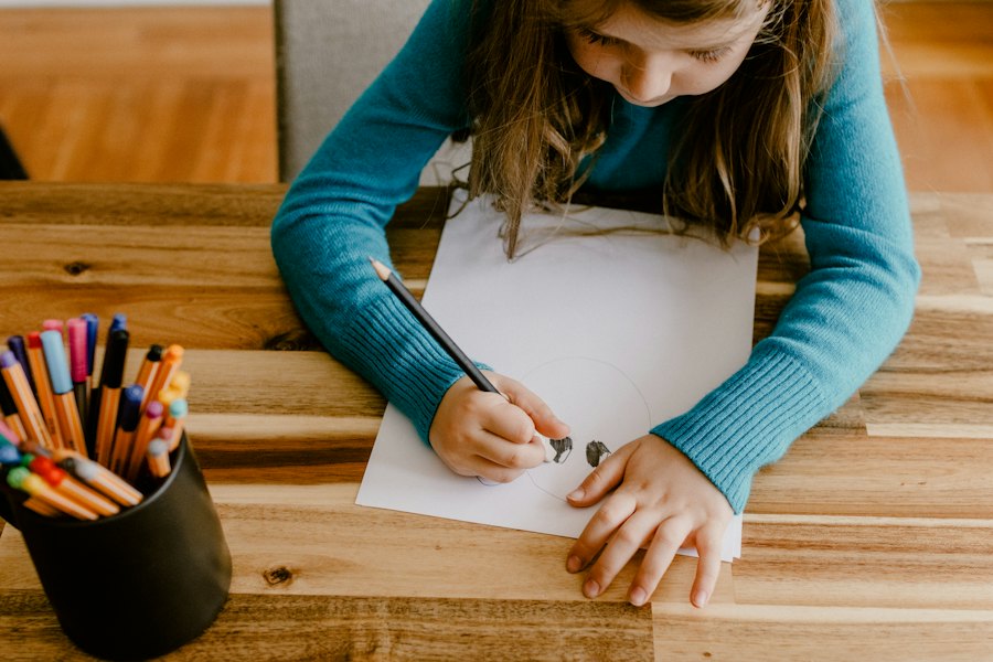 A school-age girl draws at a table next to a cup of colored pencils.